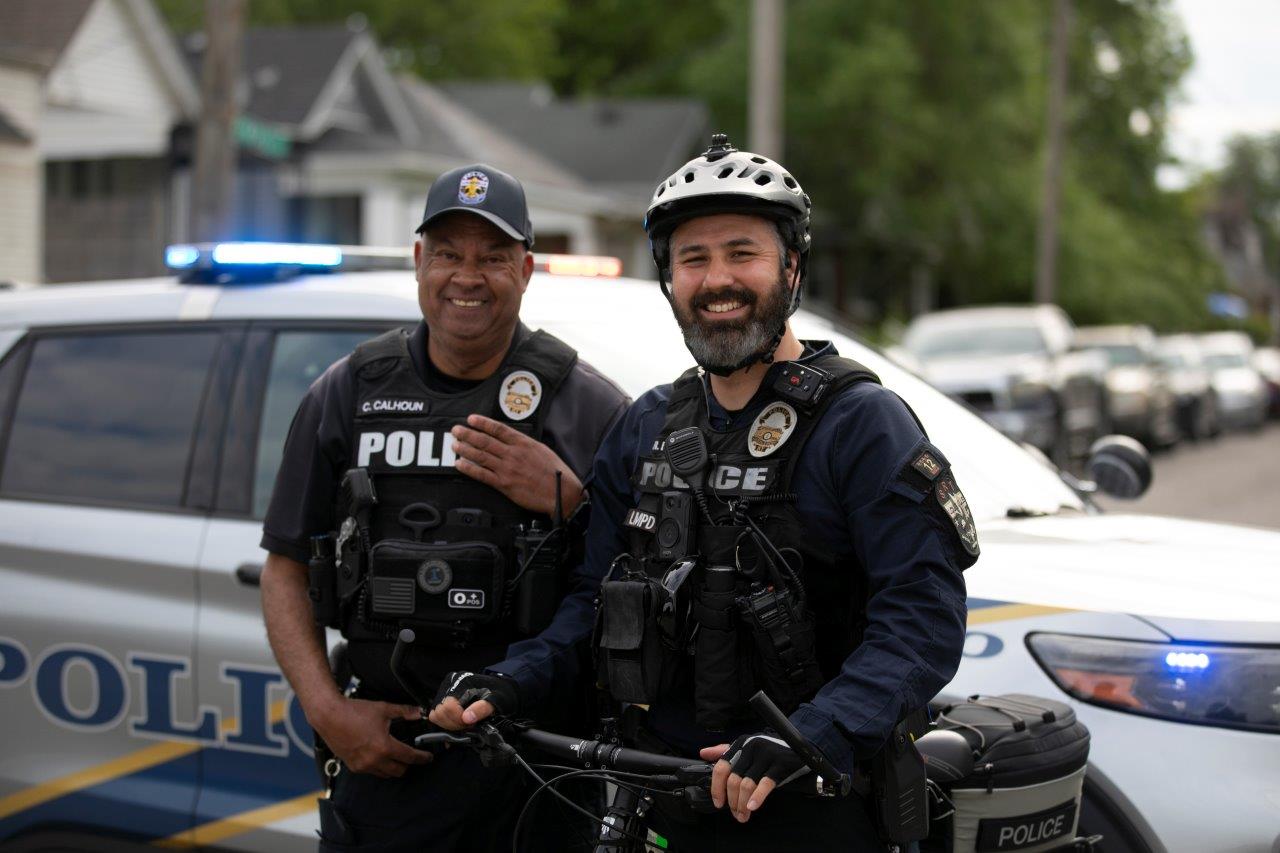 Two police officers standing in front of a police car and smiling